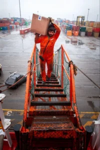 Industrial worker manually carrying a heavy load in a warehouse, showing physical strain and ergonomic risk during material handling.
